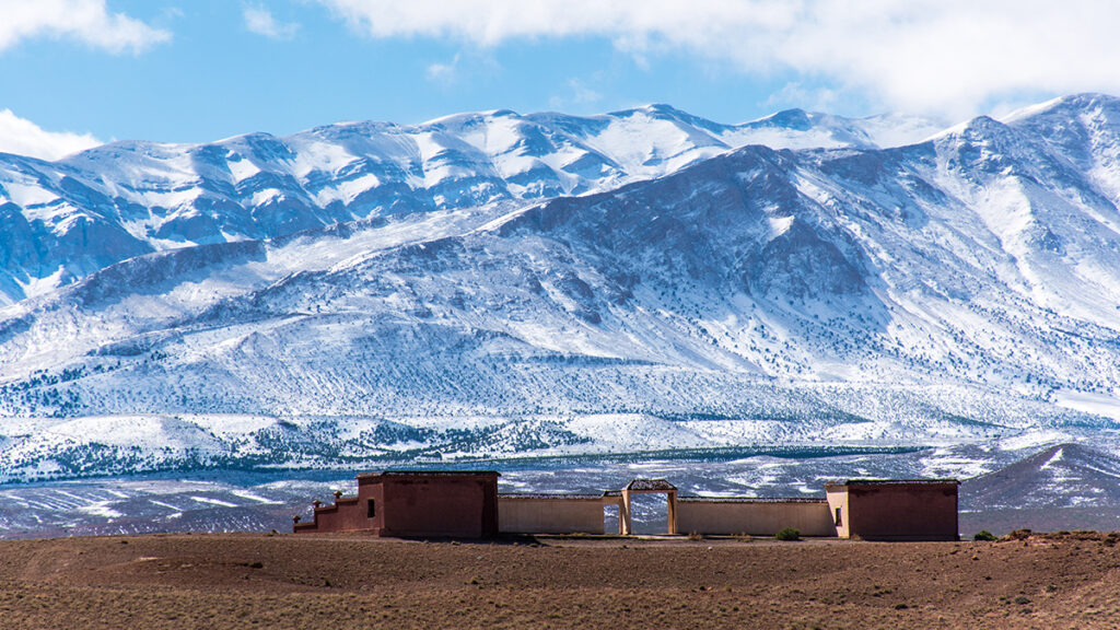 Small brown structures against a backdrop of tall, snowy mountain peaks and a blue sky