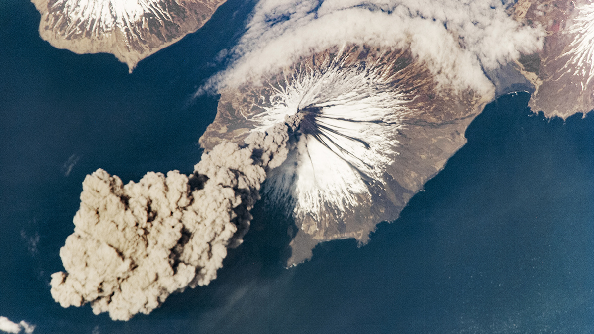 Aerial image of a gray volcano topped with snow emitting a large cloud of gray smoke. The volcano is surrounded by dark blue water.