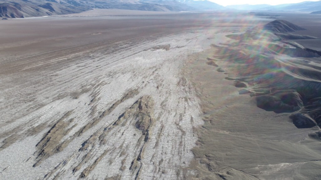 A wide, flat area of tan-colored earth, with mountains in the distance. In the foreground, a paler, gray-colored substance appears to overlay the ground. Streaks of the tan-colored ground are visible beneath the gray substance.