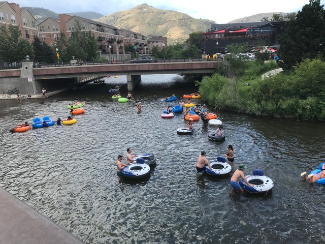A group of river tubers in the water among buildings and a bridge.