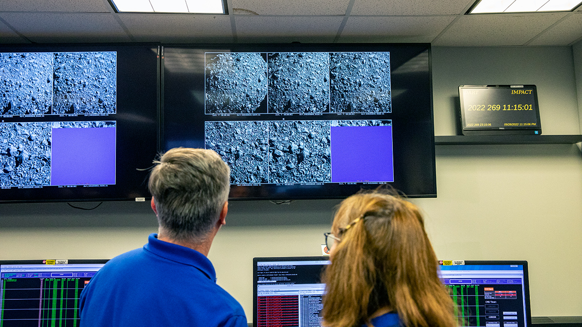 A man and a woman are seen from behind; both of them are looking at large screens showing black and white images of a rocky surface.