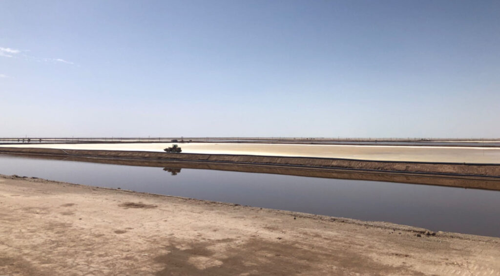 A long brine evaporation pond sits amid a bare, arid-looking landscape under a clear blue sky.