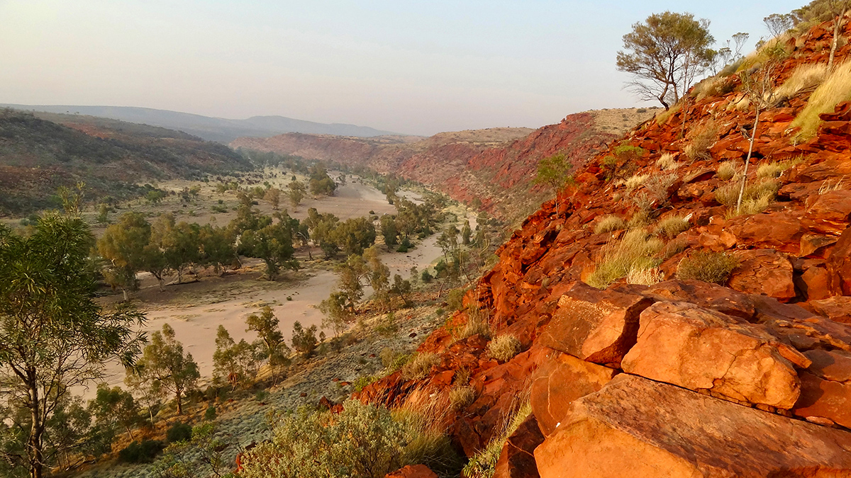 A view of a river running through a valley with green trees. On the right, in the foreground, is a ledge of red rocks.
