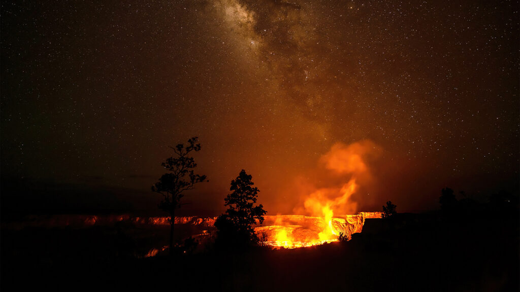 The orange glow from erupting lava illuminates the area around the summit of Kīlauea volcano under a star-filled night sky.