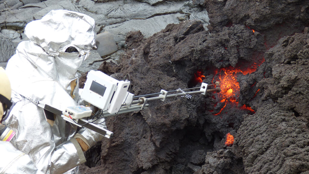 Un científico en un traje plateado inserta un tubo largo de metal en la lava incandescente naranja y en la roca gris obscuro.
