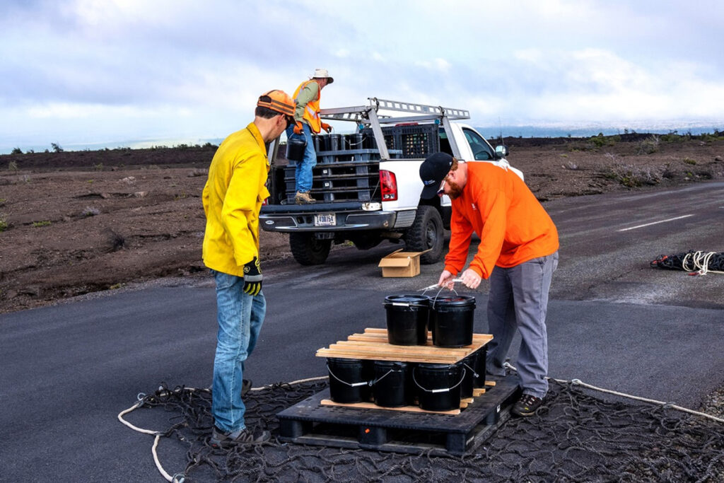Two people load black buckets onto a pallet with a large net beneath it as a third person stands in the bed of a pickup truck with more buckets and pallets.