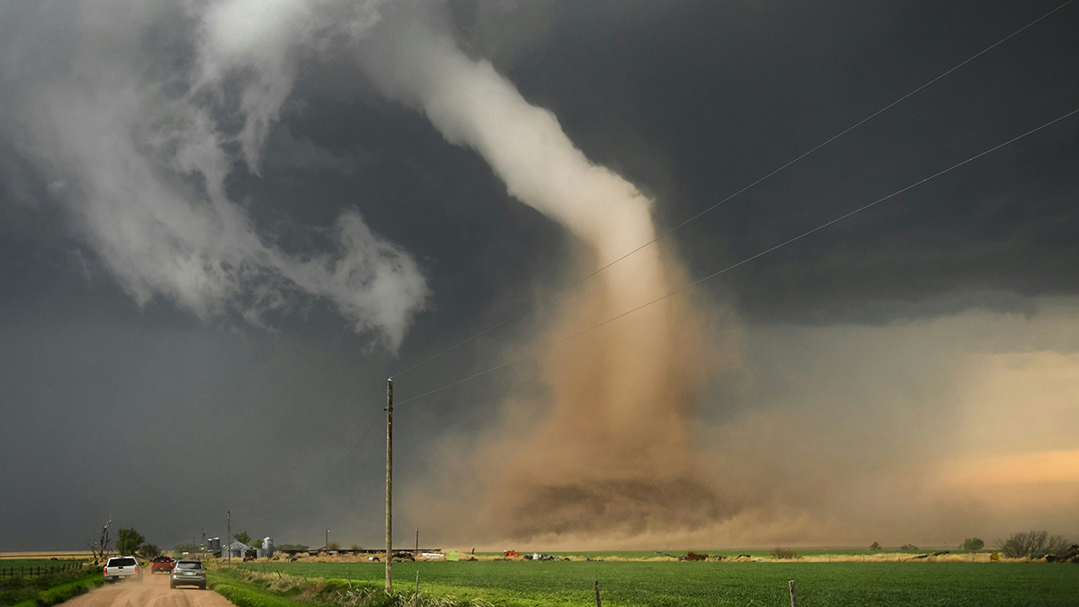 A curved tornado kicking up a brown plume of dirt beneath a dark gray sky