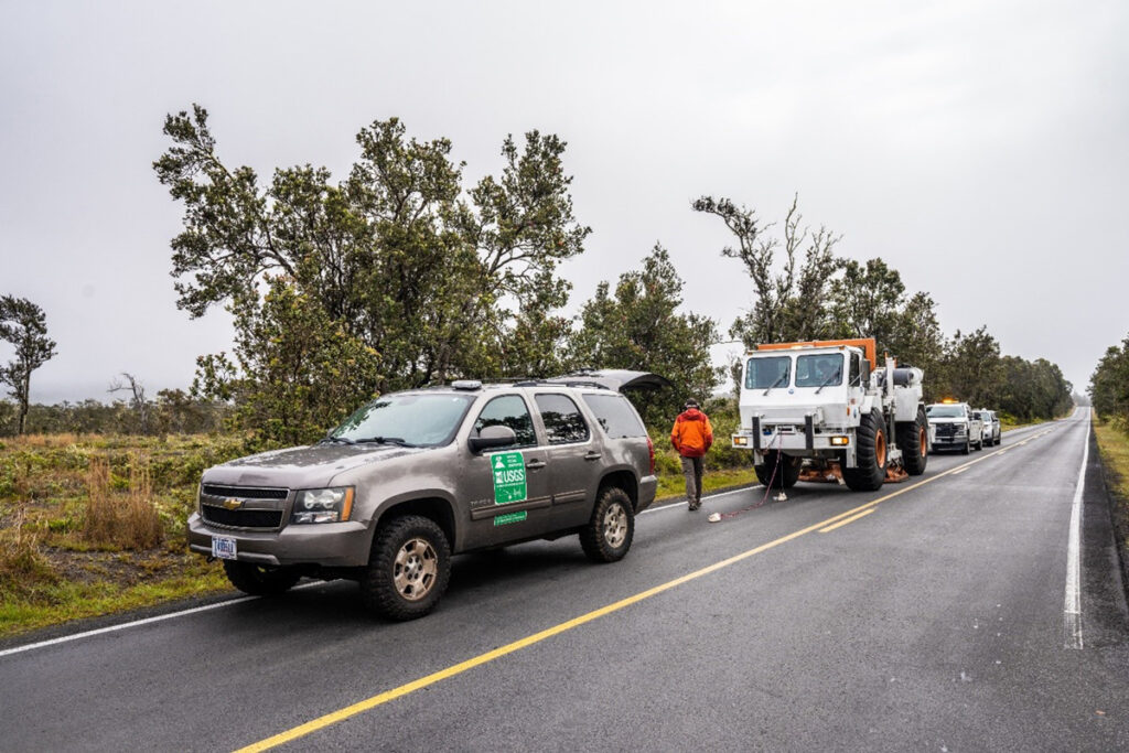 A line of four vehicles, including a U.S. Geological Survey-marked SUV and large “vibroseis” truck equipped to shake the ground, sit on a long stretch of straight roadway.