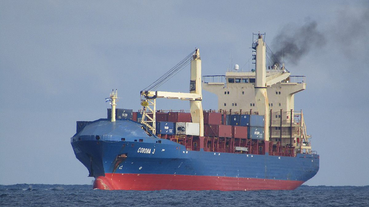 A blue and red cargo ship sails through the ocean while spewing black smoke into the air.
