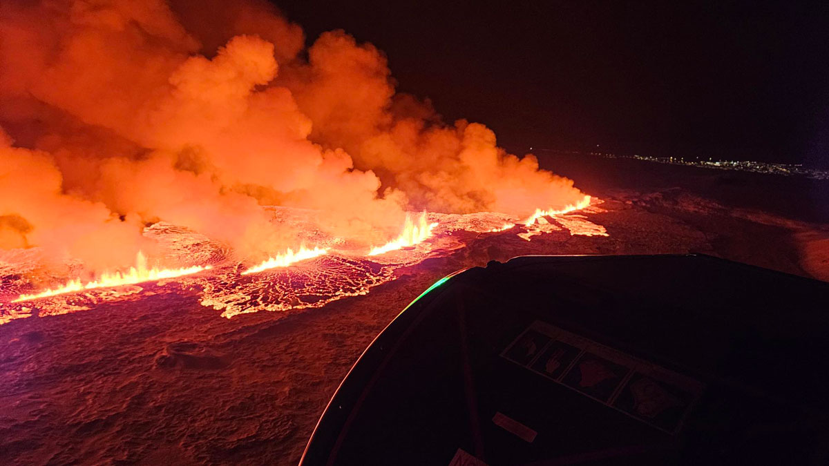 Vistas desde el aire durante la noche, varias brillantes fuentes de lava estallan a lo largo de una fisura lineal, con columnas teñidas de naranja elevándose sobre ellas. Las luces de una ciudad están a lo lejos.