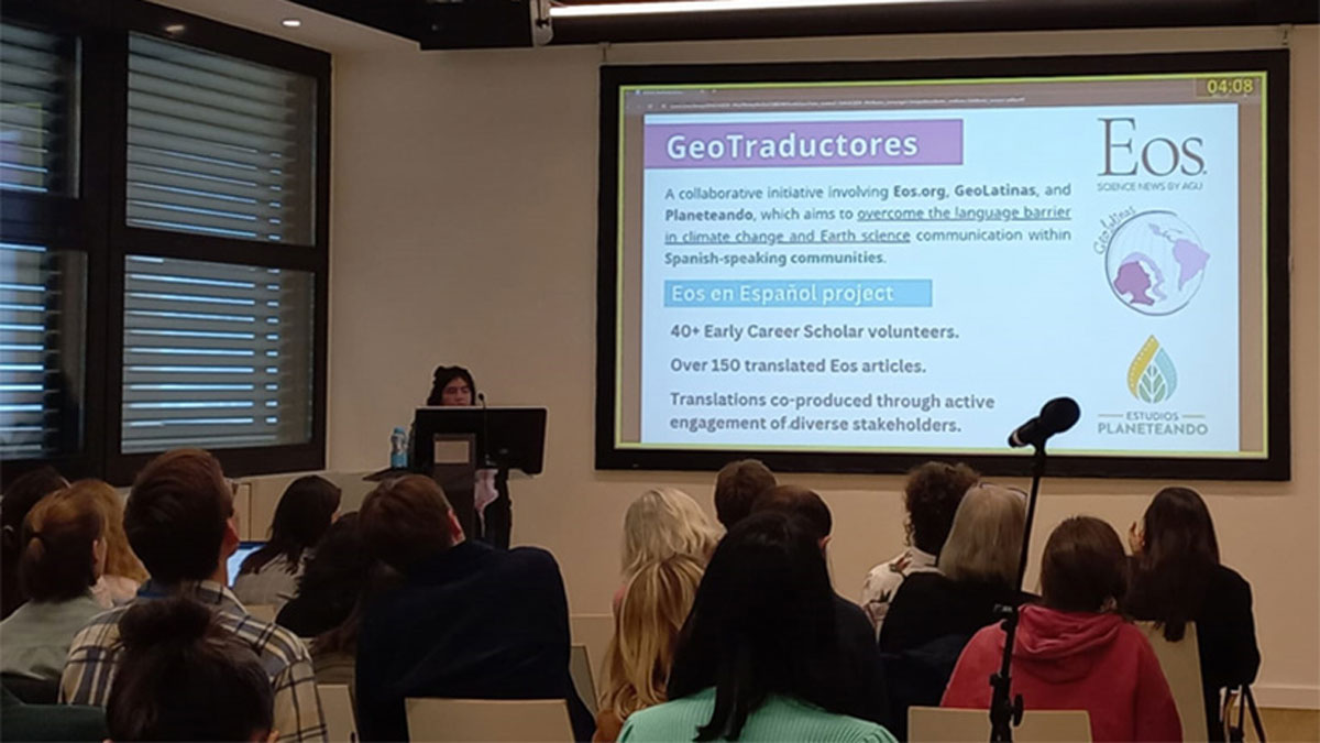 Una foto tomada desde el fondo de una pequeña sala de conferencias, donde un grupo de personas miran una mujer que está de pie en un podio y está presentando una diapositiva de Powerpoint que dice “GeoTraductores” en la parte superior.