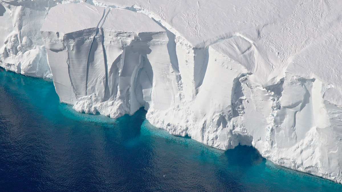 A photo of an Antarctic ice shelf in the process of calving (meaning a section is breaking off the front to become an iceberg). The water in front of the ice shelf is a deep blue.
