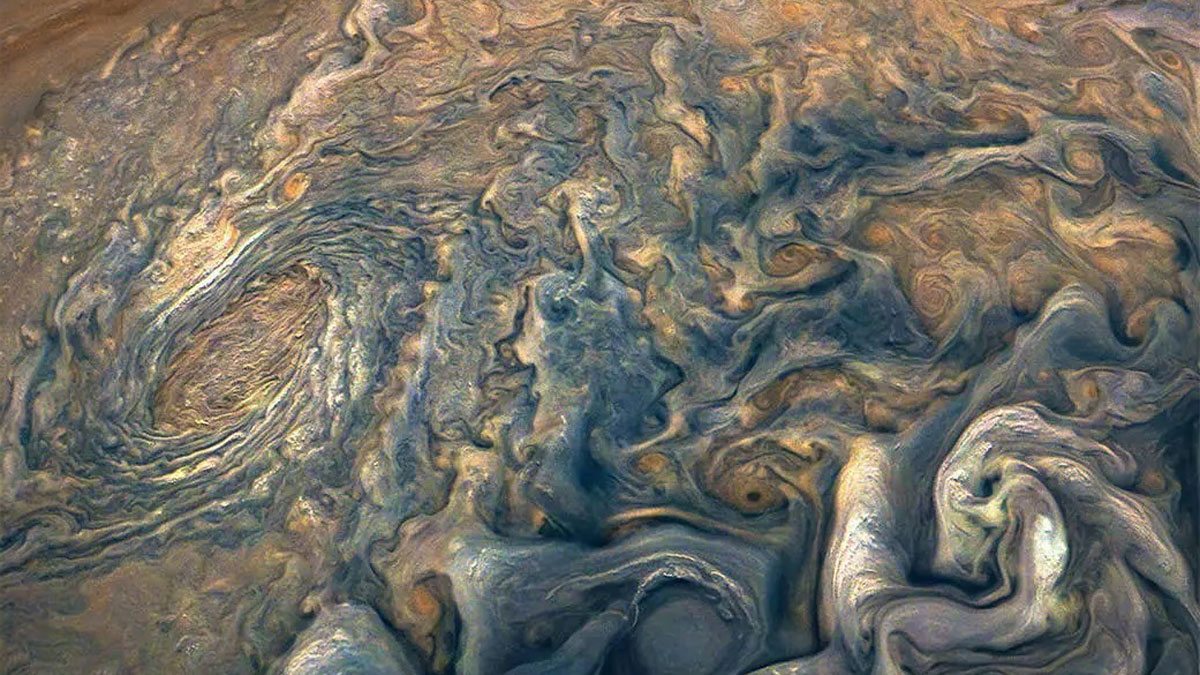 An image of Jupiter’s atmosphere, which is full of striking, swirling, blue and light orange clouds.