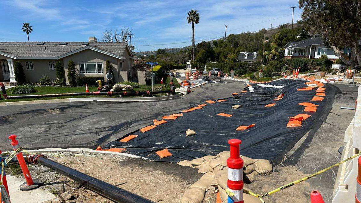 A road runs through a neighborhood; the road is warped and covered with black tarp and orange sandbags in some places.