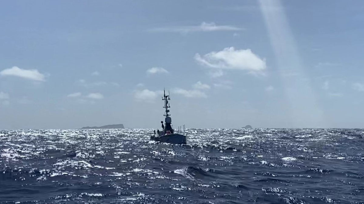 A ship traveling on the ocean is seen in silhouette. The sky is blue with some wispy clouds, and the sea is reflecting lots of sunshine.