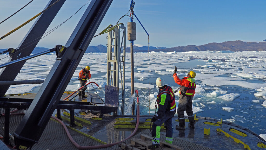 Three people wearing orange safety jackets stand on the deck of a ship in the Arctic.