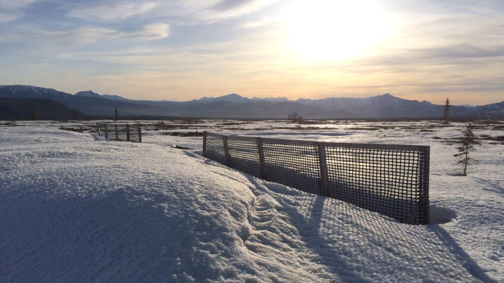 Two short stretches of fencing beside piled snow amid an open stretch of land, with mountains in the distance.