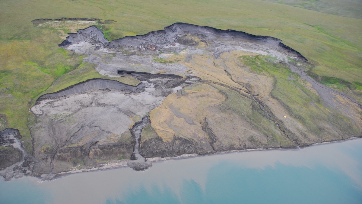 A coastline is seen from above. The area is mostly covered in greenery, but what appears to be a large landslide has uncovered gray soil that is falling into the blue ocean.