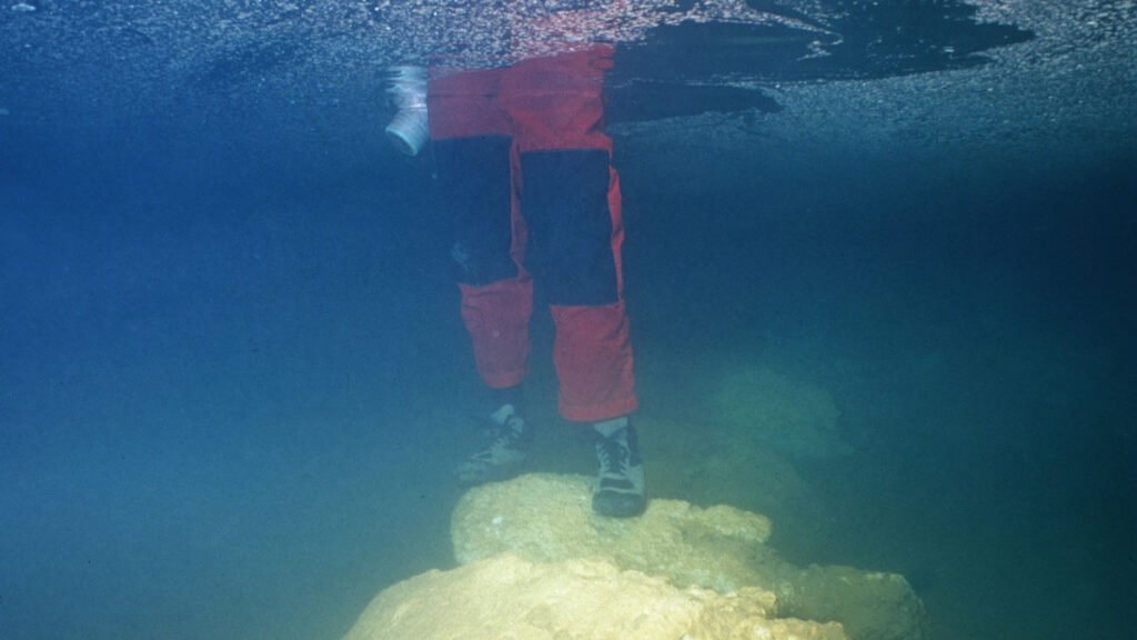 An underwater picture of a scientist wearing red and black waterproof pants and boots standing in thigh-high water on an ancient underwater bridge.