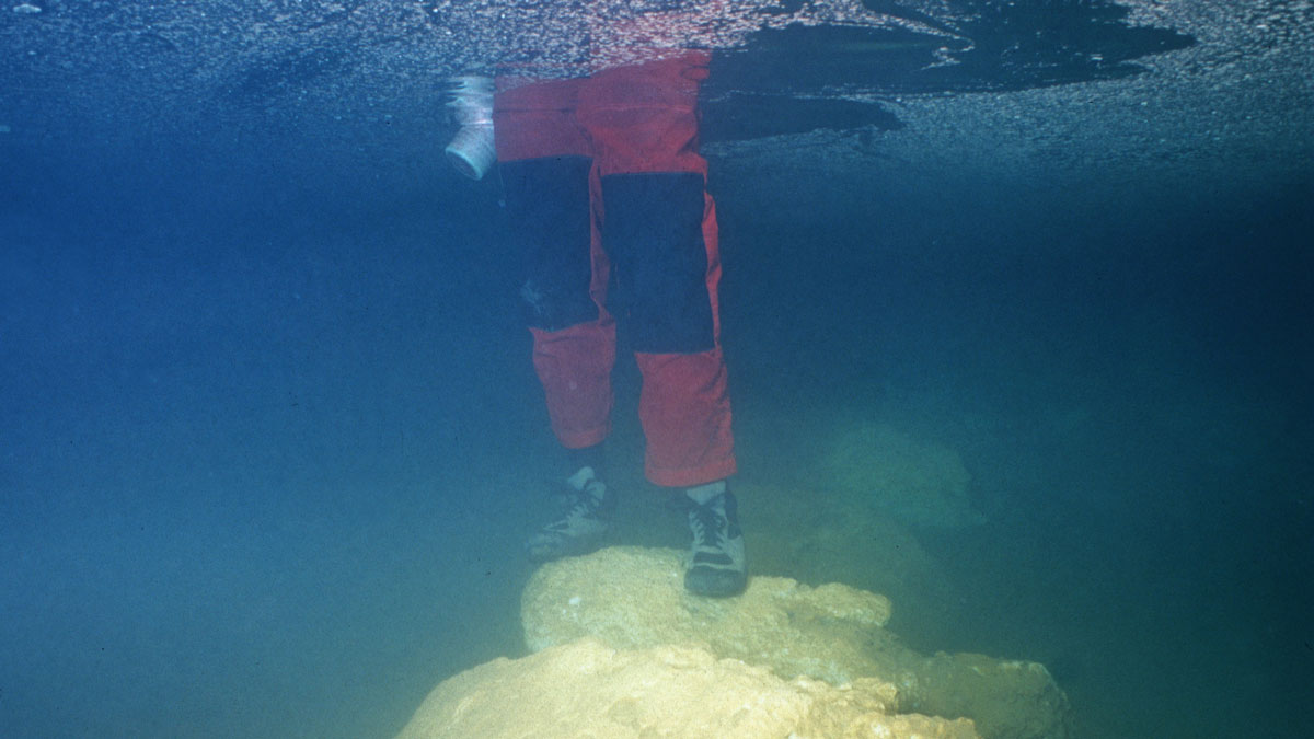 An underwater picture of a scientist wearing red and black waterproof pants and boots standing in thigh-high water on an ancient underwater bridge.