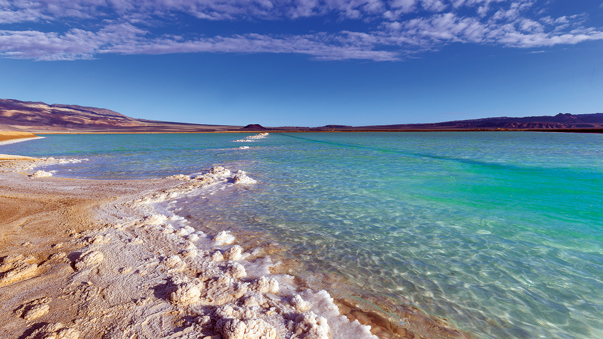 An aquamarine body of water in a desert landscape. The water’s edge has white crystallized material.