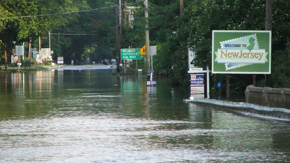 Floodwater over a road. A sign reads, “Welcome to New Jersey.”