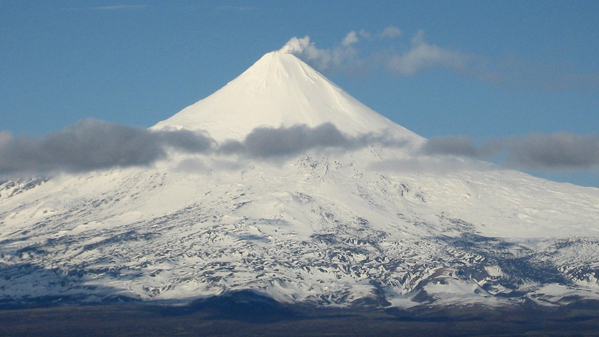 The top of a conical volcano covered in snow appears against a blue sky