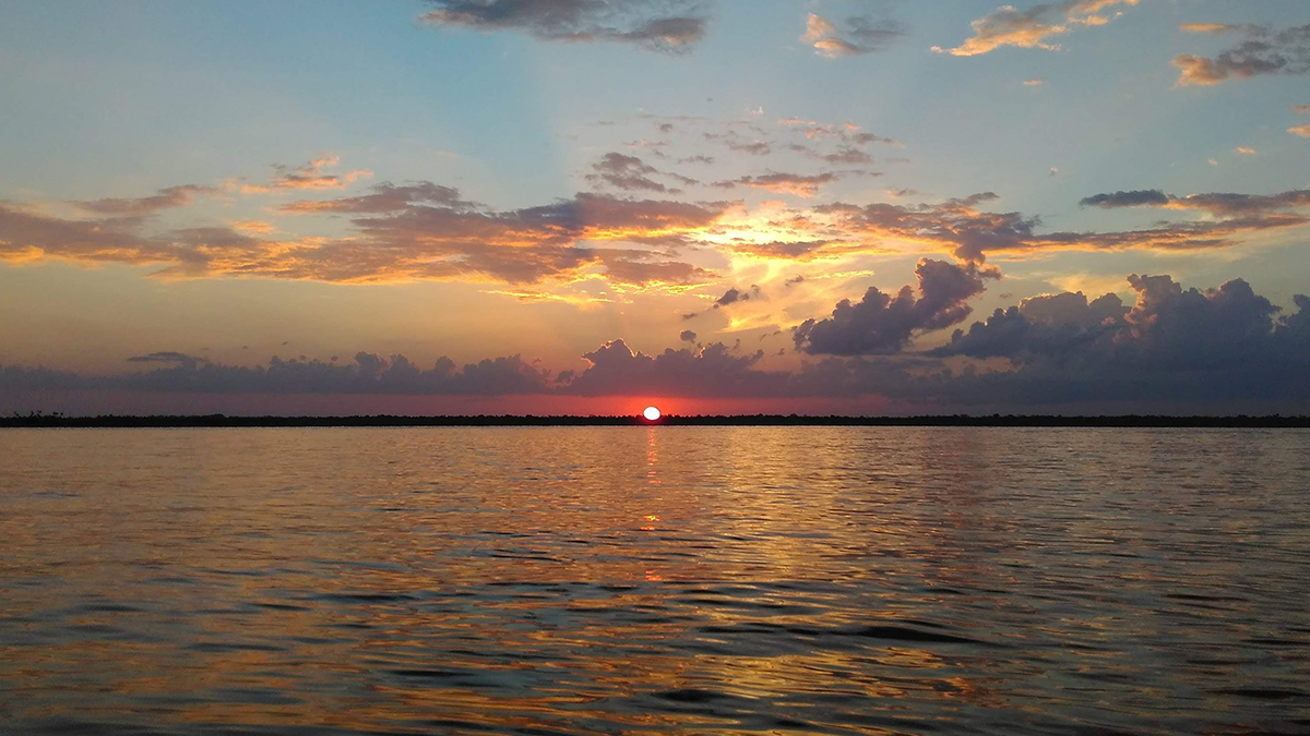 O Sol poente fica logo acima do horizonte e sob um céu parcialmente nublado, com um corpo de água em primeiro plano.