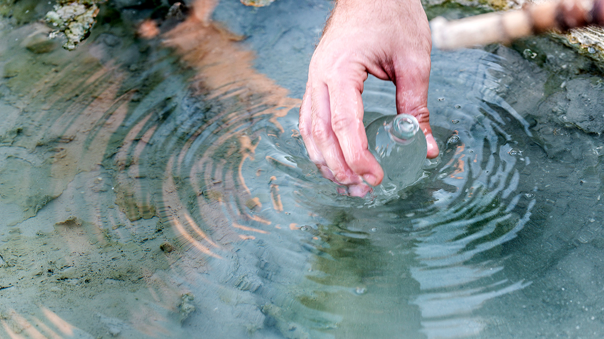 A person’s hand is shown dipping a sampling bottle into a shallow pool of water.