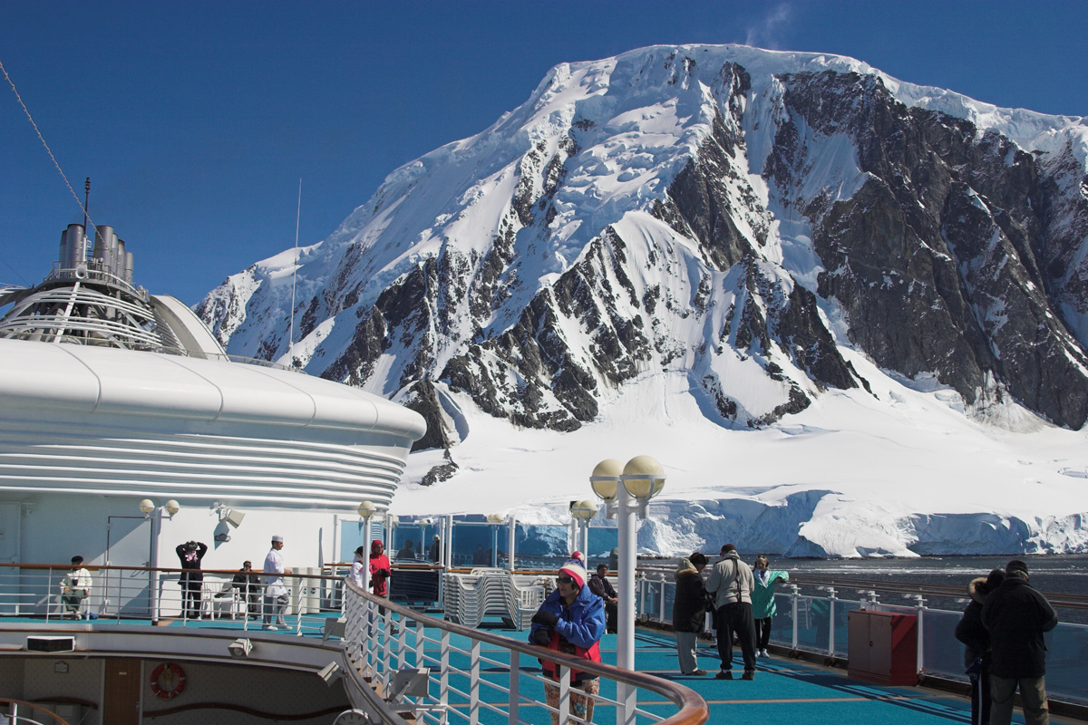 A photo taken from the deck of a cruise ship in Antarctica, featuring people bundled in jackets looking up at a snowy mountain.