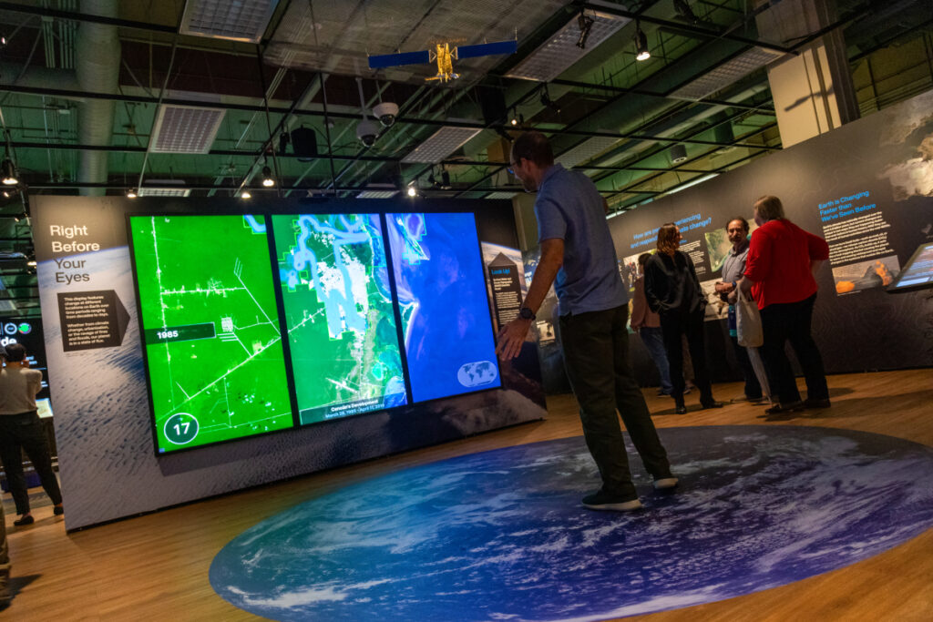 : A person stands on a floor decal of Earth in front of three digital displays. An outline of the person’s body appears over the data.