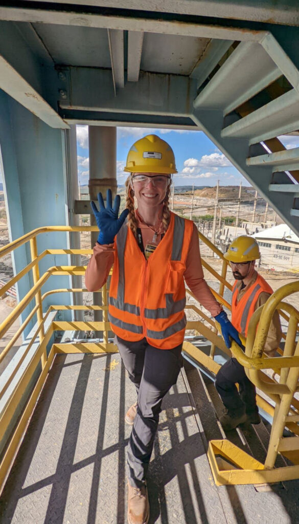 A young woman in an orange vest, safety goggles, and a hard hat waves at the camera. She is on an outdoor stair landing.