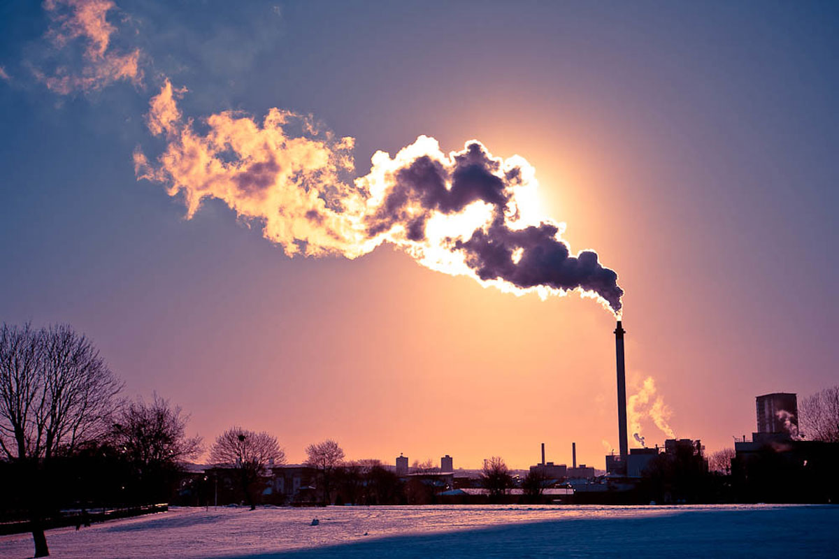A smokestack emits smoke, which blocks out the Sun and is lit up against a blue and orange sky.