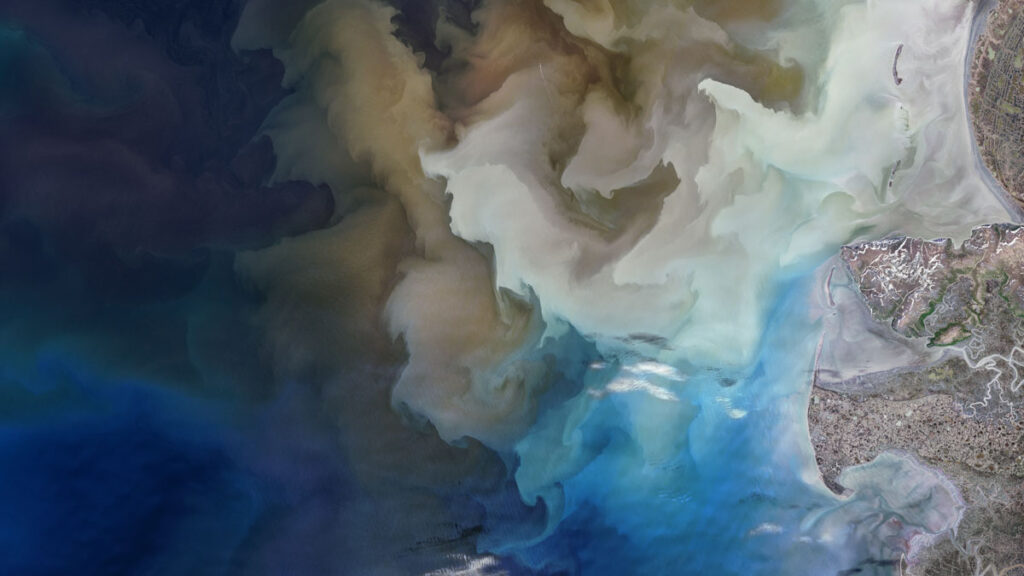 Satellite imagery of Alaska’s coastline. Fans of gray and brown silt appear stacked on top of each other in the blue ocean.