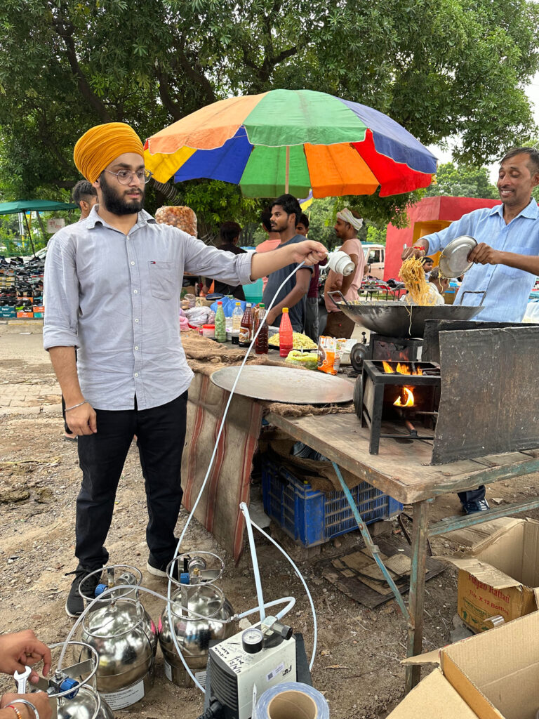 A scientist holds an emissions sampling device above a lit kerosene stove sitting on a table at an outdoor food stand.