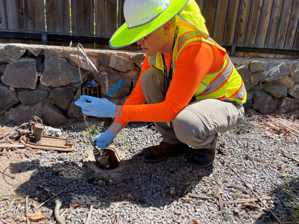 A person in an orange top and yellow reflective vest and hat crouches in front of an outdoor tap to collect a water sample in a brown glass bottle.