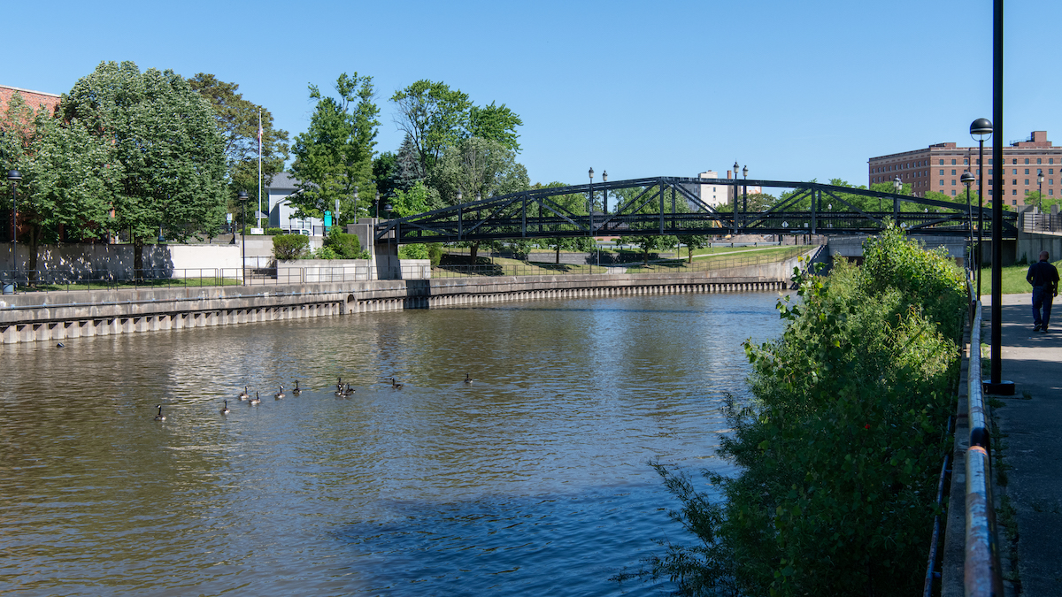 A river in an urban setting flows under a black metal bridge on a sunny day.