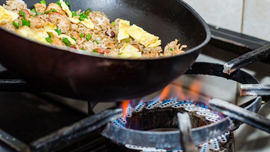 A rice and vegetable dish is being cooked in a wok over a lit gas burner.