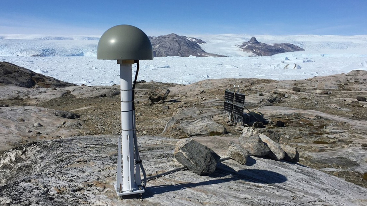 A metal pole with a small gray-green dome, anchored to bare rock. Glacial ice is visible in the background.