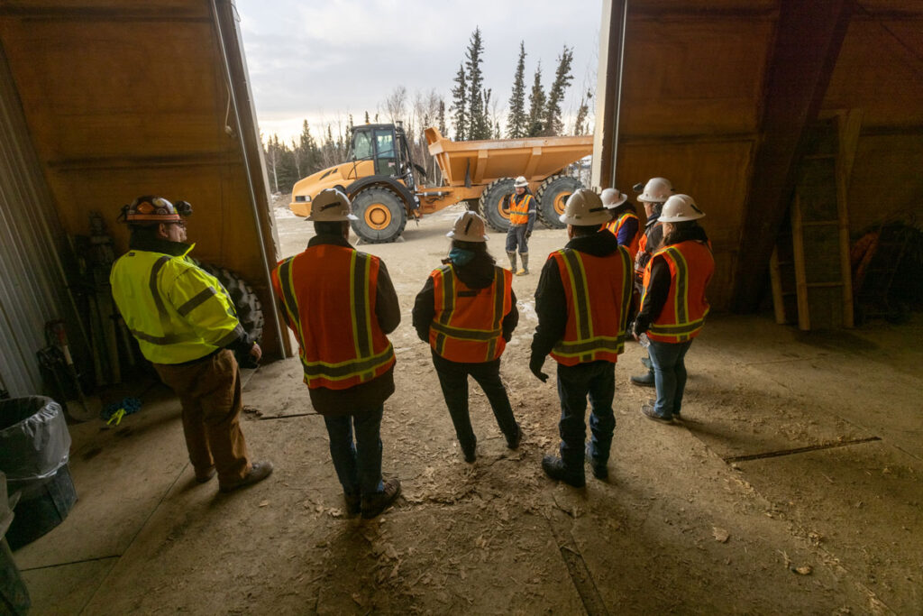 A group of people in hard hats and bright orange stand inside a building, looking out through a large door at a dump truck.