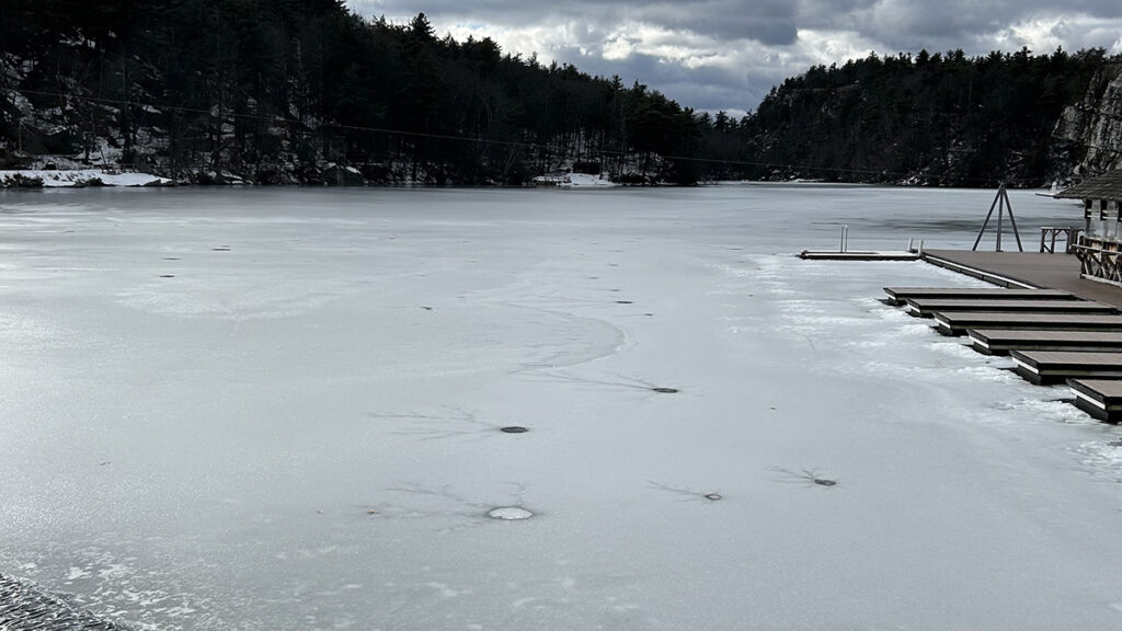 A lake partly covered in ice can be seen with mountains and clouds in the background. A dock is visible on the right side of the photo.