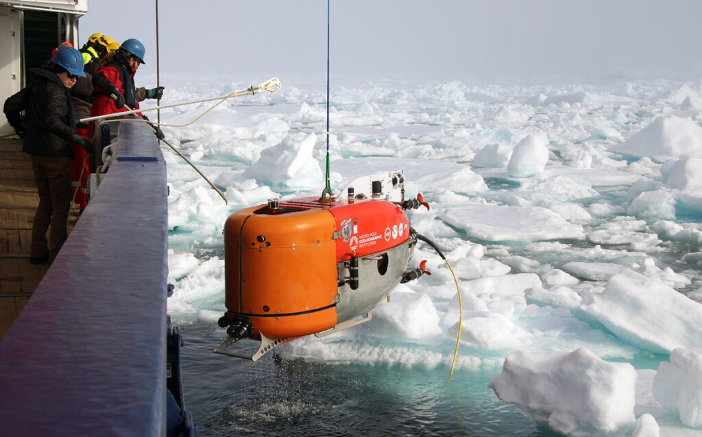 Scientists lean over the side of a ship, reaching for a submersible suspended from a rope, which has just emerged from icy waters.