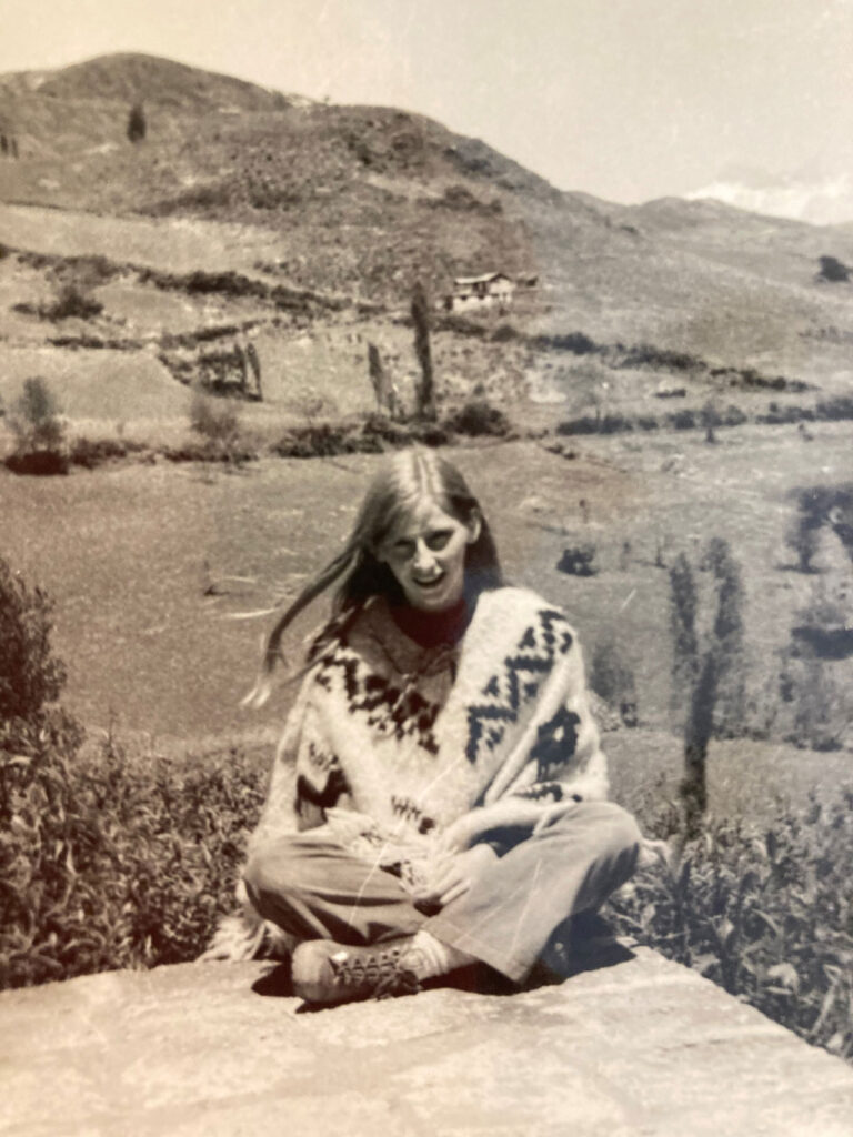 Black-and-white photo of a young woman sitting cross-legged on a rock. There are mountains in the background.