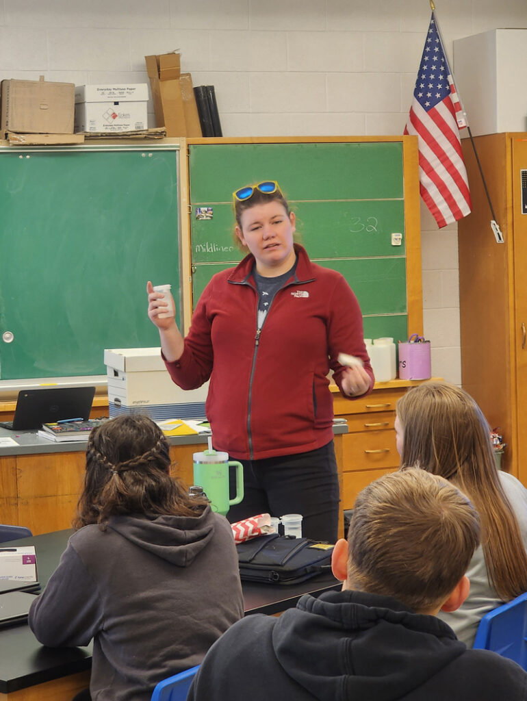 A woman holding a test tube of water speaks to a classroom of high school students, who are seen from behind.