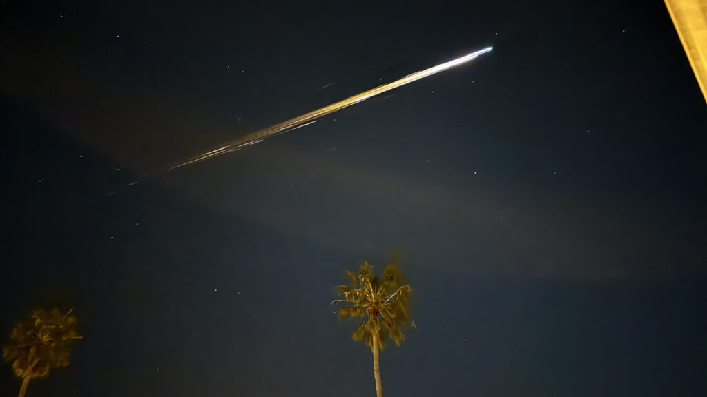 View of a starry night sky with streaks of white and yellow through the center. Front-lit palm trees appear in the foreground.