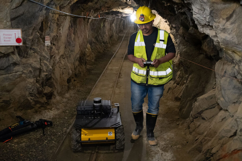 A person in a hard hat and bright yellow vest stands in a mine, remotely operating two toy-sized trucks.