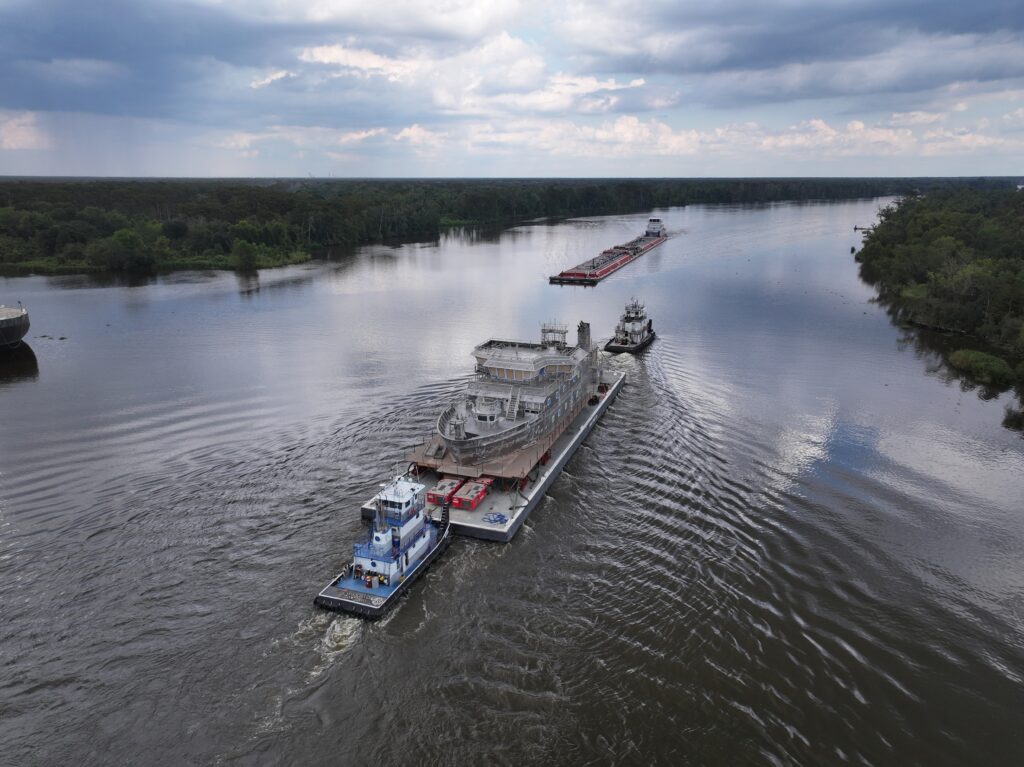 Two tugboats move a larger, unfinished ship structure down a river.