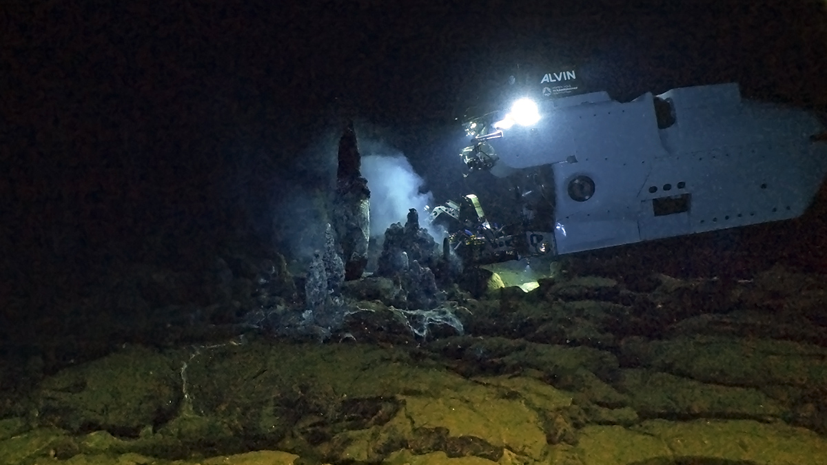 The Alvin submersible hovers over and illuminates the seafloor near the rocky towers of a hydrothermal vent site.