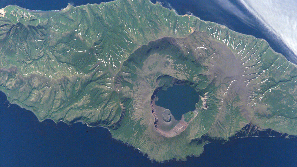 A top-down view of green and brown land with a roughly circular depression with a blue lake in the middle