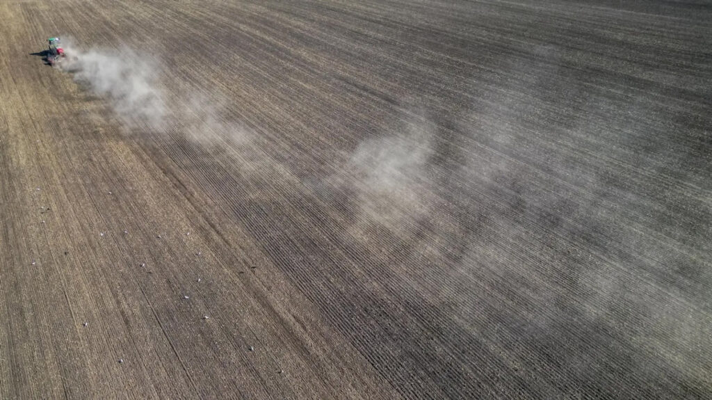An overhead view of a farmer driving a vehicle through a field, leaving behind a trail of billowing dust